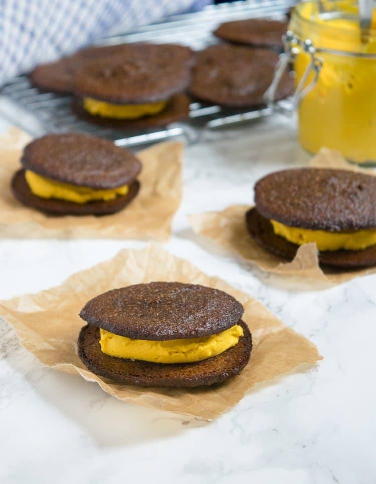 Pumpkin whoopie pies with pumpkin cream filling on squares of parchment paper, with more cookies on a cooling rack behind