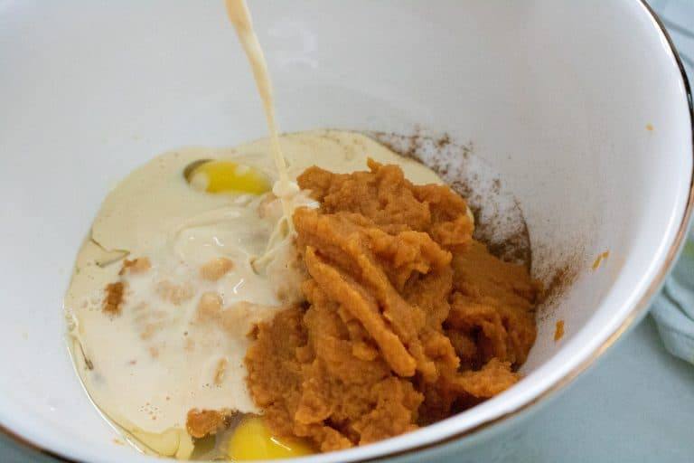 Evaporated milk being poured over pumpkin puree and eggs in a bowl of spices for pumpkin meringue pie
