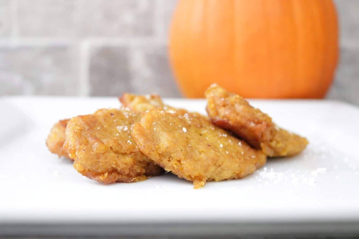 Golden pumpkin patties dusted with powdered sugar on a white plate with a whole pumpkin in the background