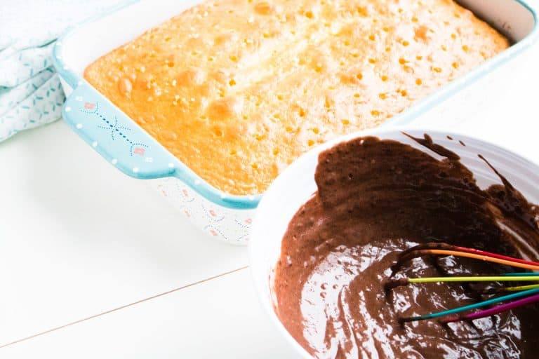 baked white cake for quadruple chocolate poke cake in 13x9 glass pan with white mixing bowl with chocolate pudding filling and whisk