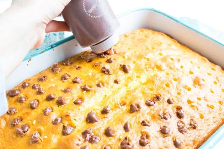 baked white cake for quadruple chocolate poke cake in 13x9 glass pan and candy decorating bottle shown filling holes poked into cake