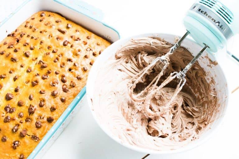 baked white cake for quadruple chocolate poke cake in 13x9 glass pan with holes filled with chocolate filling with white mixing bowl with cream cheese and chocolate frosting in it with blue mixer to the side