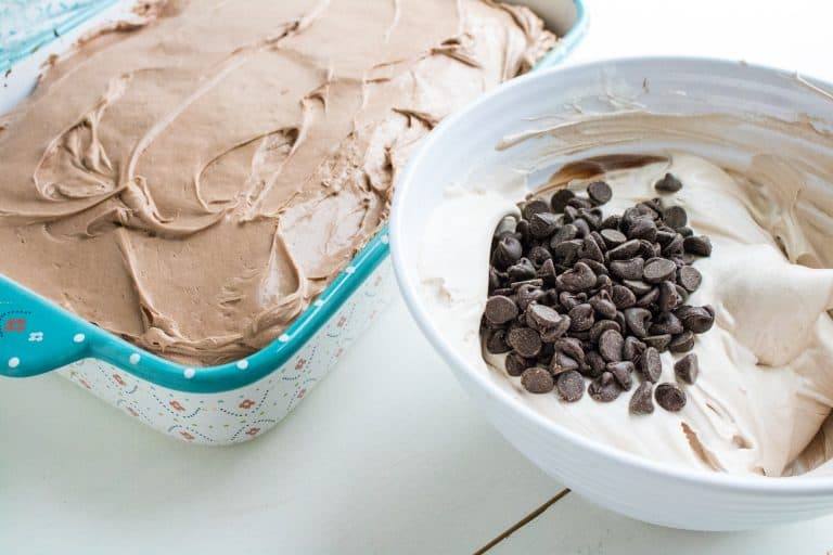frosted cake for quadruple chocolate poke cake in 13x9 glass floral pan with white mixing bowl next to it with whipped topping with chocolate chips on top waiting to be mixed in