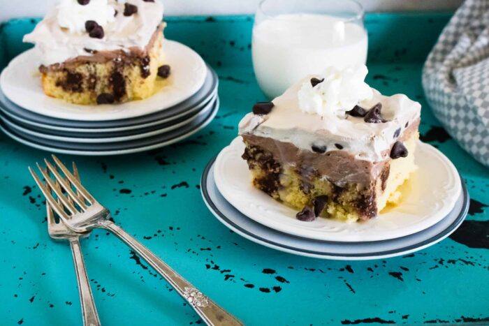 Two slices of Quadruple Chocolate Poke Cake served on white plates set on top of a blue tablecloth