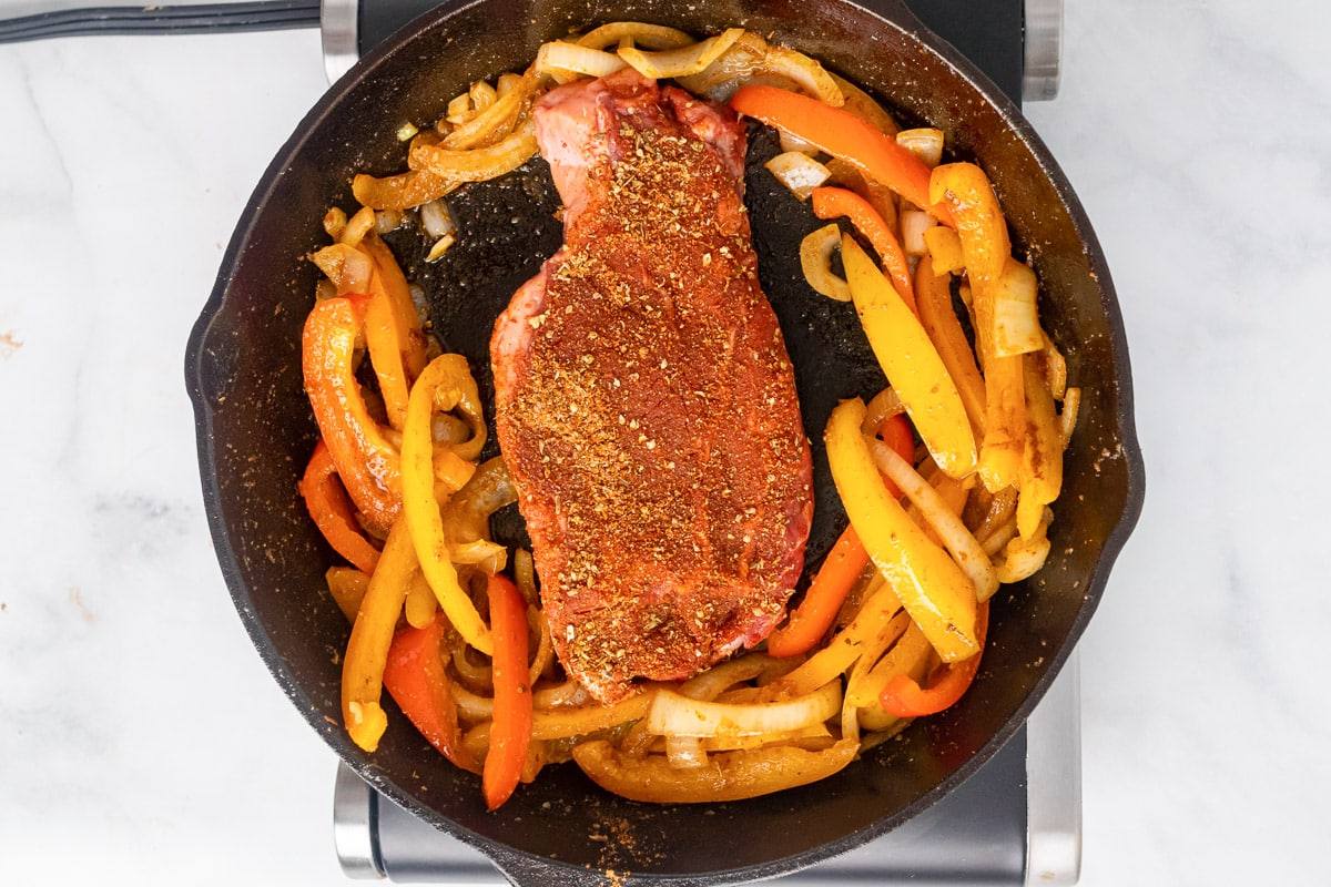 seasoned steak in skillet with cooked peppers and onions ready to be pan fried