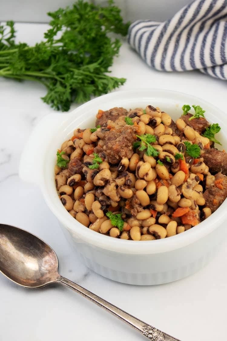 Black eyed peas with beef stew meat and carrots in a white serving dish with a spoon and fresh parsley