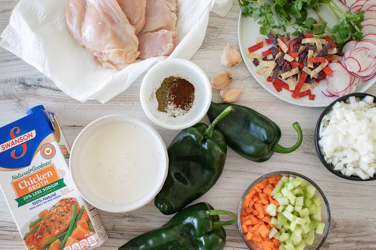 Ingredients for chicken poblano soup: raw chicken breasts, poblano peppers, diced onion, carrots and celery, chicken broth, heavy cream and spices