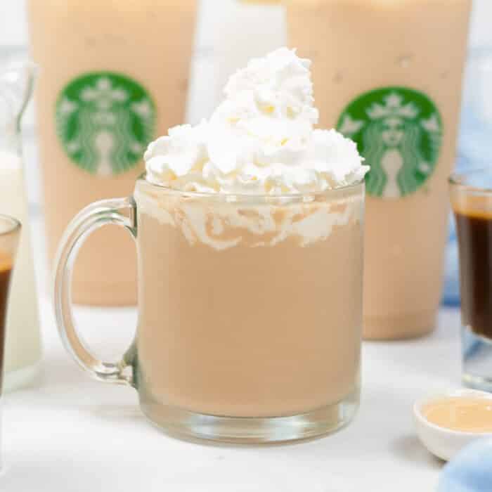 copycat starbucks white chocolate mocha in a clear glass mug topped with whipped cream in front of two iced Starbucks cups