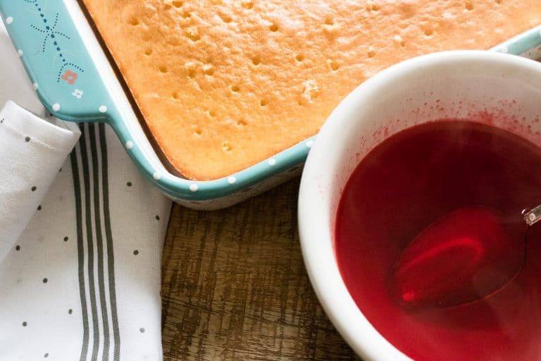 baked white sheet cake with holes poked all over the top, sitting next to a white bowl of the liquid strawberry gelatin mixture