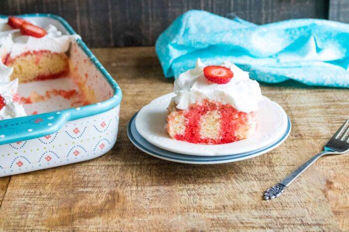 Completed Strawberry Poke Cake in a blue print sheet pan sitting next to a slice served on a white dessert plate with a blue rim