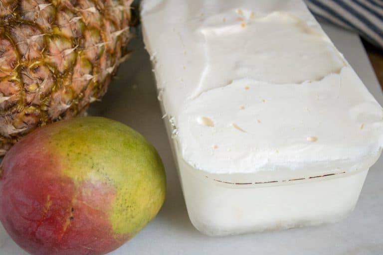 a container filled with tropical no-churn ice cream sitting next to a whole, uncut mango and pineapple