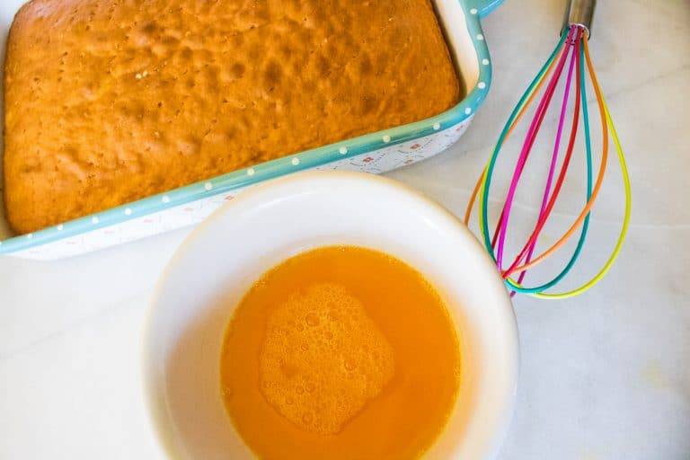 Top down view of baked white cake in a sheet pan with holes poked all over the top of the cake, next to a white bowl filled with mango gelatin liquid and a rainbow colored wisk