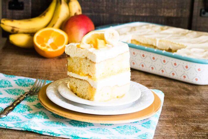 Slice of Tropical Poke Cake serves on a white and orange dessert plate set on top of a blue print napkin with a silver fork