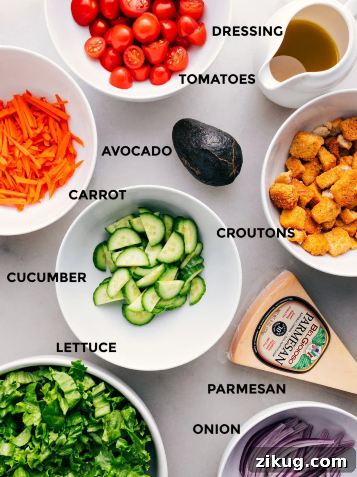 Essential fresh ingredients for a garden salad laid out: crisp lettuce, ripe avocado, Parmesan cheese, crunchy croutons, refreshing cucumbers, juicy tomatoes, and a small bowl of dressing.