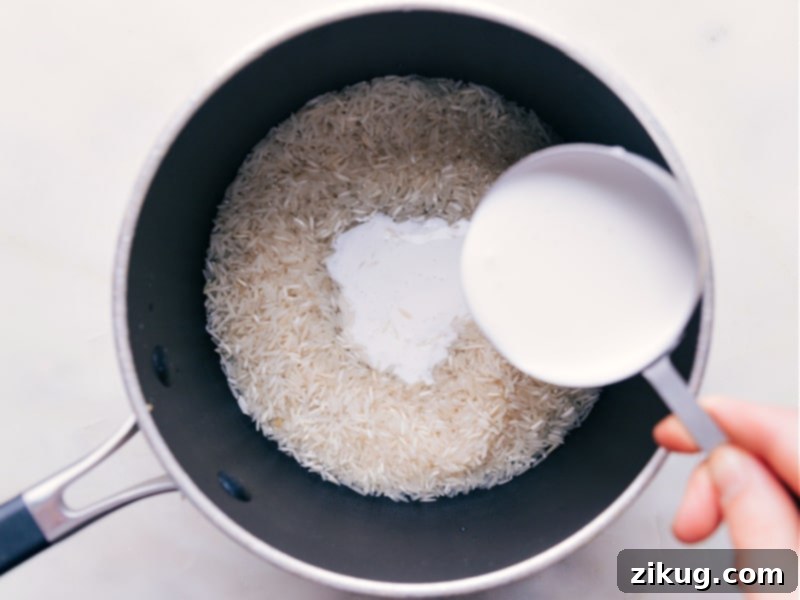 Coconut Rice is such a fun switch-up from typical rice -- it makes a great side dish or a fantastic base to so many dishes! It has a lovely, yet subtle, coconut flavor with a hint of freshness from the lime. Recipe via ChelseasMessyApron #dinner #side #best #quick #easy #simple #lime #kidfriendly #familyfriendly #coconut #rice Pouring creamy full-fat coconut milk from a can into a pot with Basmati rice and water, preparing to cook the delicious coconut rice. The scene highlights the essential liquid ingredient.