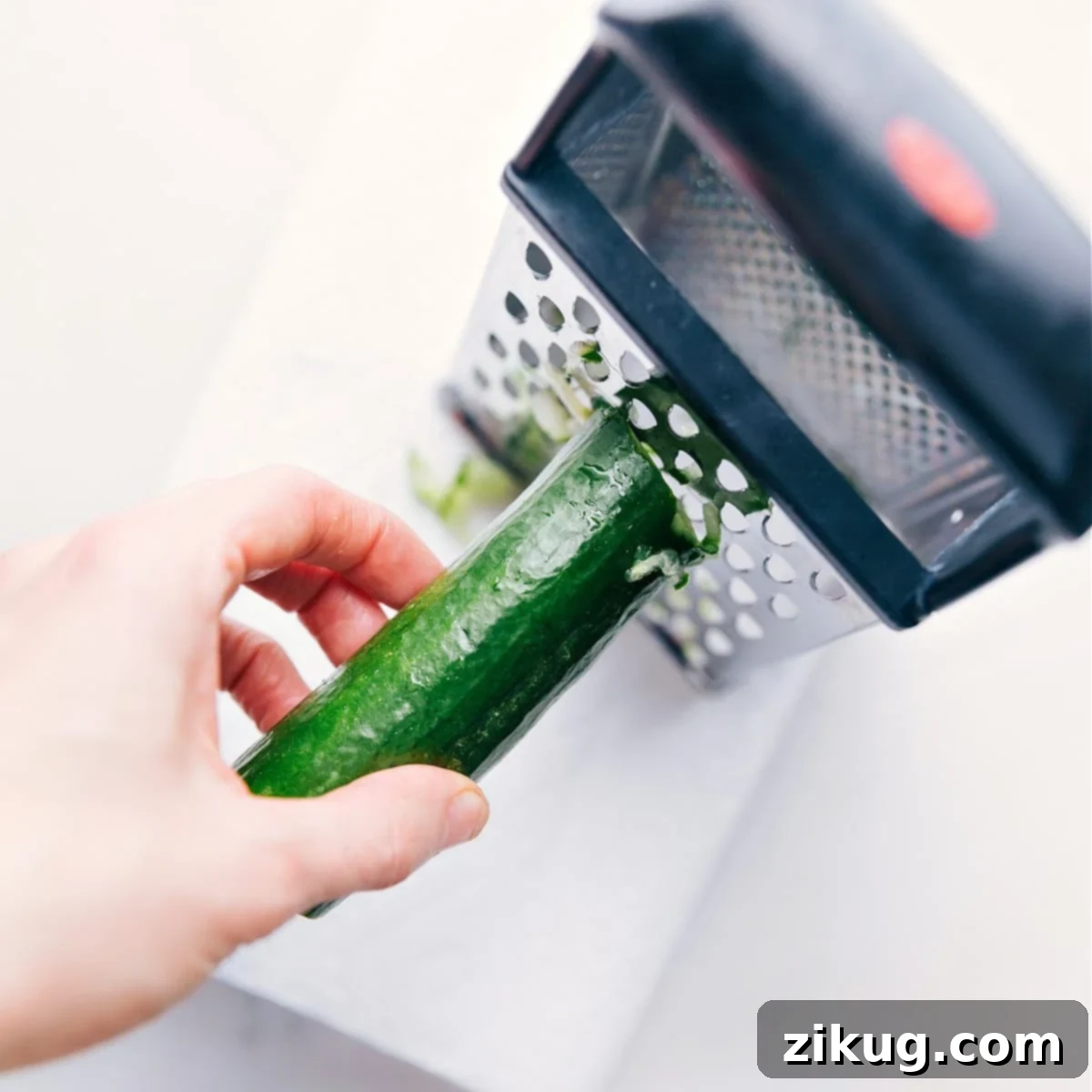 Grating Cucumber for the Freshest Tzatziki Sauce A close-up shot of hands grating an English cucumber directly into a bowl using a box grater, highlighting the initial step of preparing fresh Tzatziki Sauce.