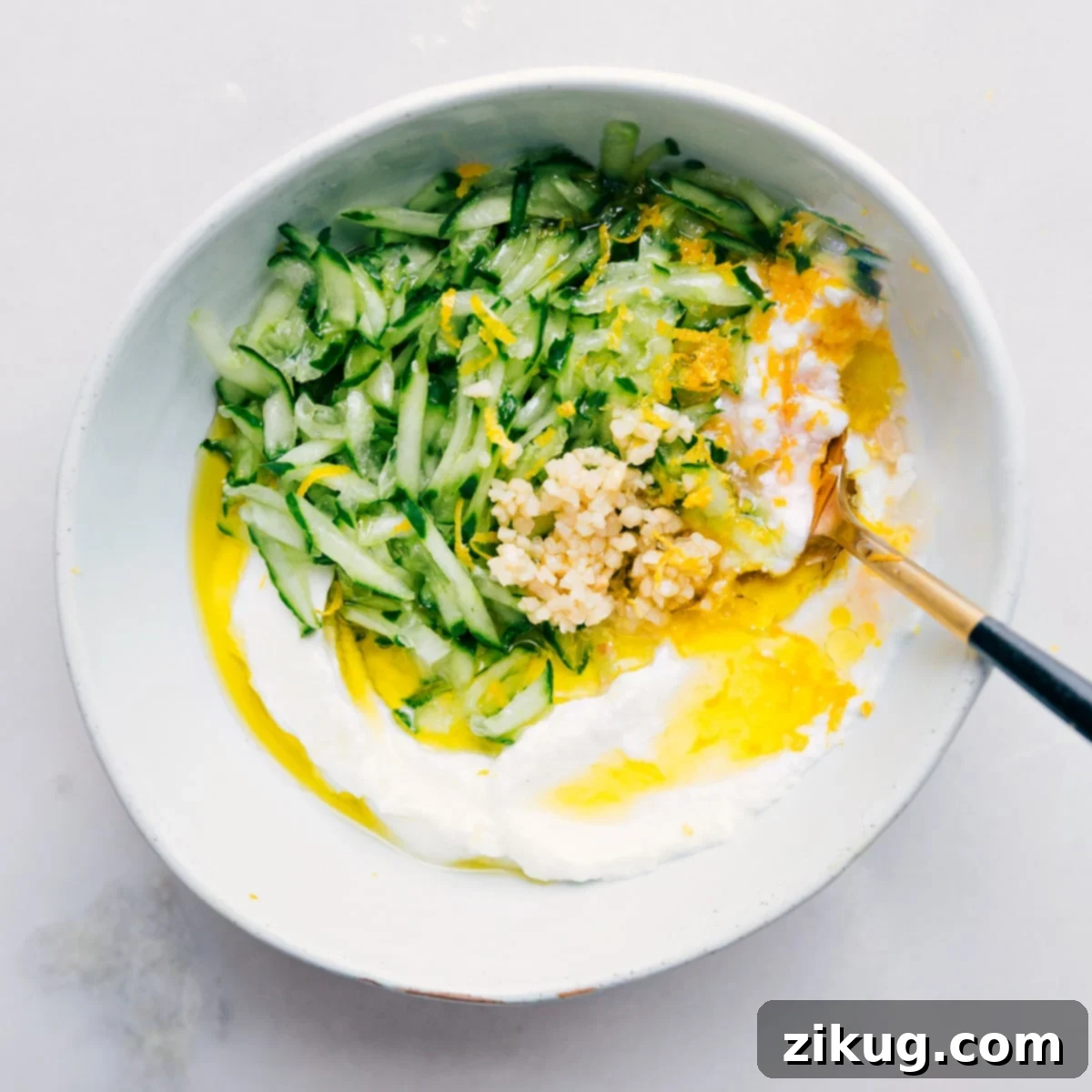 Effortless Mixing: All Your Tzatziki Ingredients in One Bowl All the freshly prepped Tzatziki ingredients, including thoroughly drained cucumber, creamy Greek yogurt, vibrant lemon zest and juice, minced garlic, and aromatic fresh herbs, being combined and mixed together in a large glass bowl.
