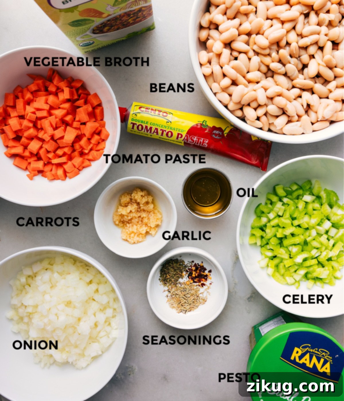 All the ingredients for this recipe are prepped and ready for easy assembly, including broth, beans, paste, oil, garlic, veggies, seasonings, and pesto, neatly arranged on a counter.