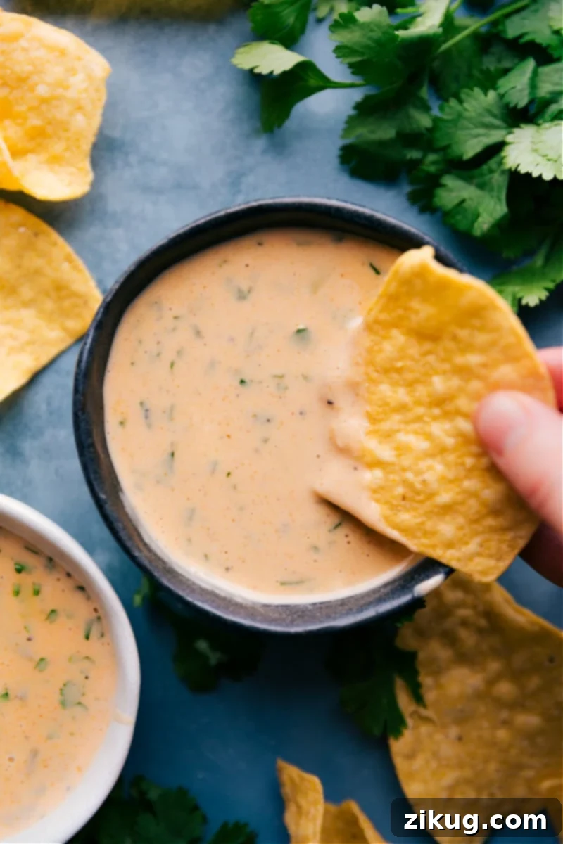 The ultimate close-up of our perfect, ultra-creamy Queso Dip, glistening and ready to be scooped up by salty tortilla chips for an unforgettable bite, a guaranteed crowd-pleaser for any event. Up-close overhead image of the perfectly smooth and creamy Queso Dip recipe, served warm in a rustic bowl, garnished with fresh cilantro, ready to be enjoyed with crispy tortilla chips.