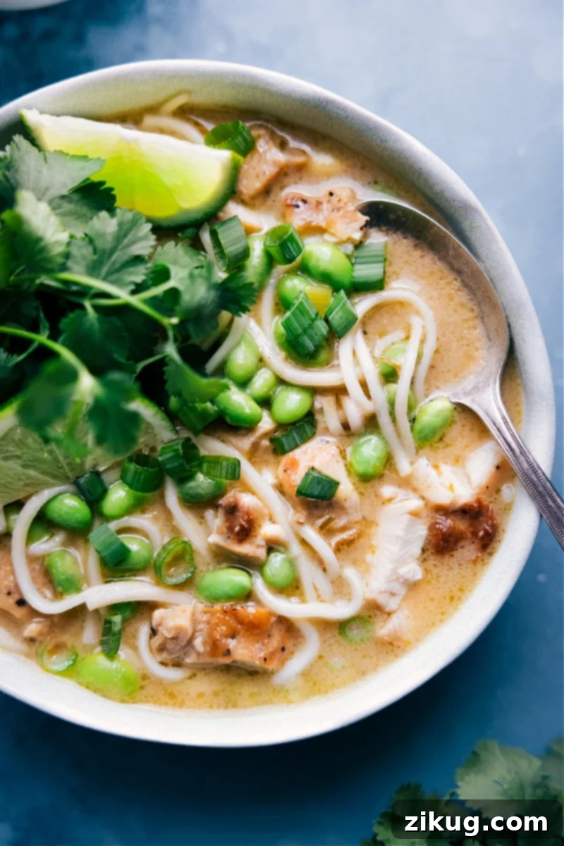 A steaming bowl of vibrant Green Curry Noodle Soup, filled with chicken, noodles, and edamame, garnished with fresh cilantro and lime wedges.