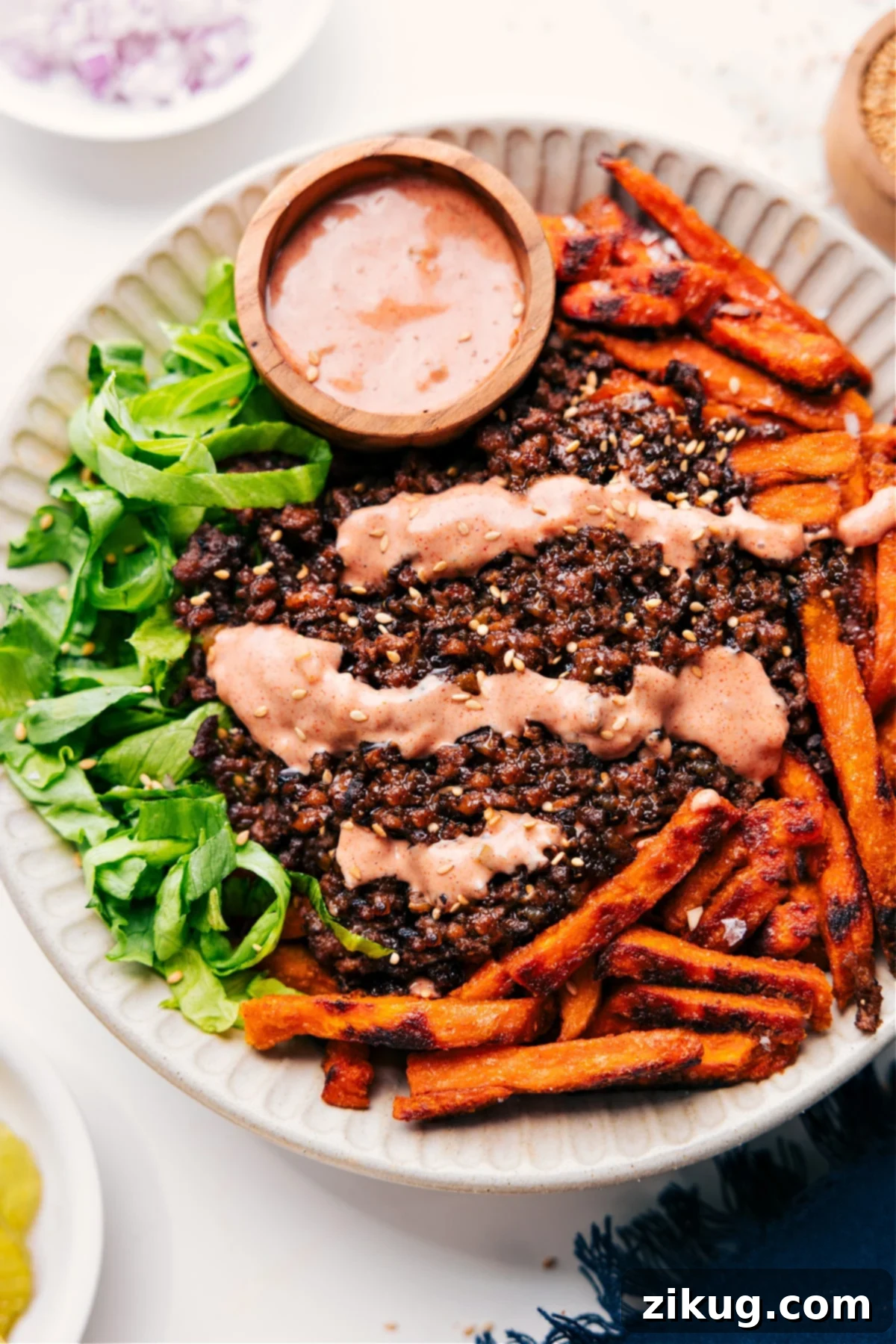 A vibrant Burger Bowl overflowing with crisp lettuce, golden sweet potato fries, savory ground beef, drizzled generously with a special burger sauce and sprinkled with sesame seeds.