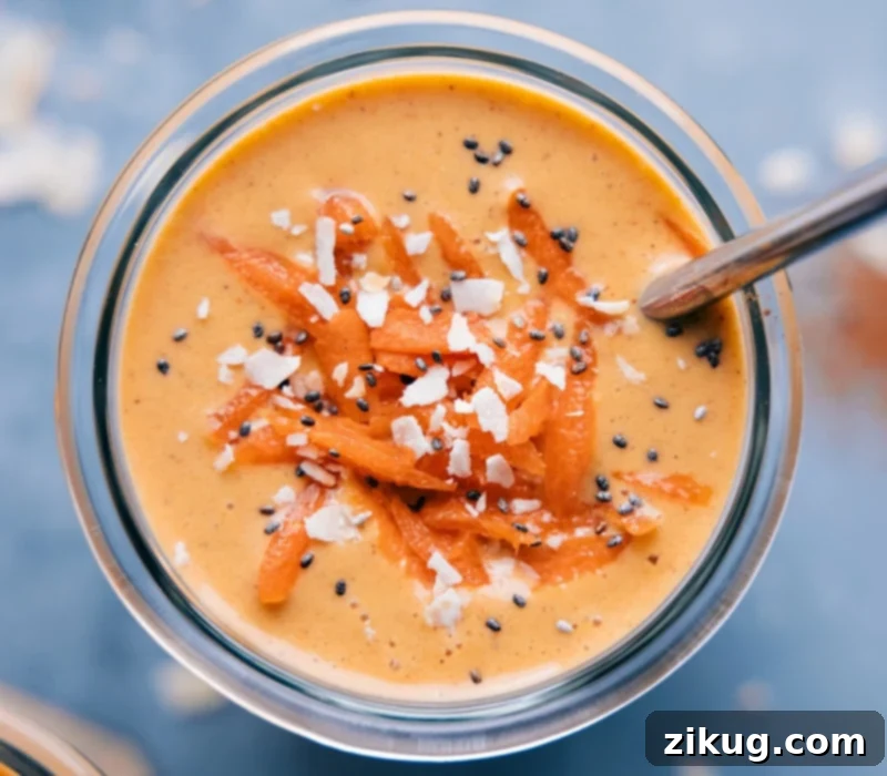 This insanely yummy Carrot Smoothie is the perfect way to channel your cravings for Carrot Cake without having to bake anything. This protein-packed, nutritious, and filling smoothie is healthful, but somehow tastes almost as indulgent as a cake! Recipe via ChelseasMessyApron #quick #easy #simple #snack #best #healthy #fresh #cleaneating #wholefoods #carrot #smoothie A beautifully presented Carrot Smoothie in a clear glass, topped with fresh shredded coconut, a sprinkle of chia seeds, and a few grated carrot strands for garnish. A straw is placed in the glass, inviting a sip.