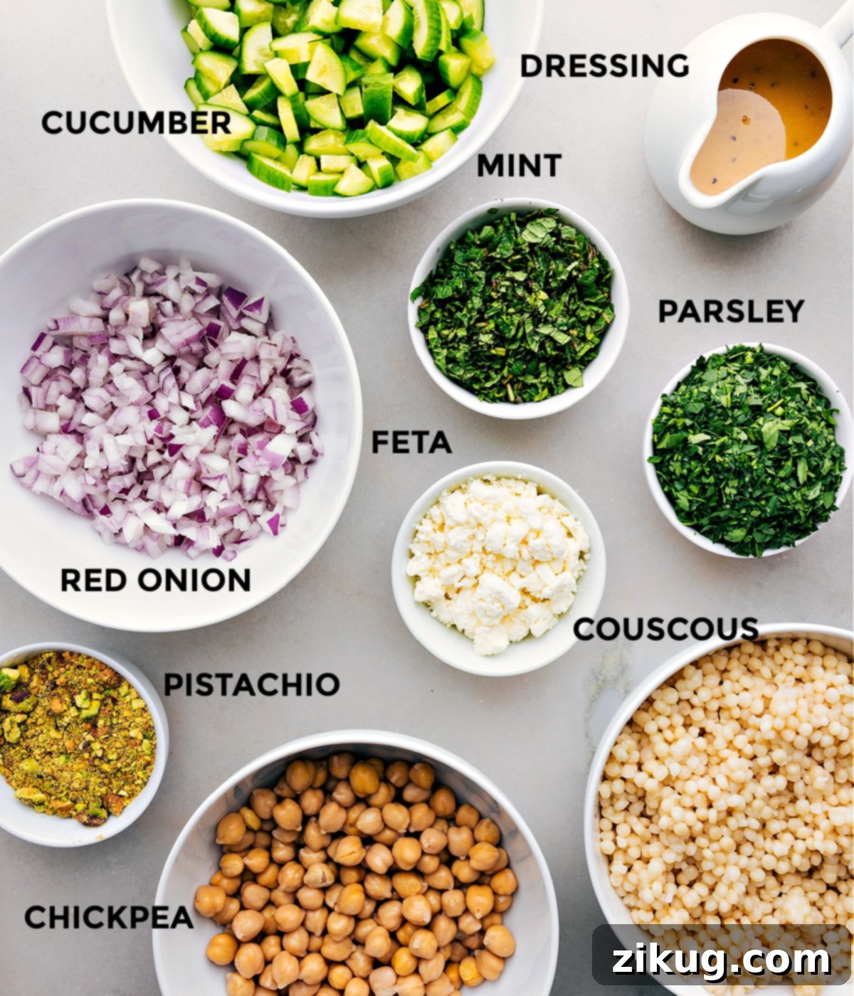 Ingredients In This Delicious Jennifer Aniston Salad for Health-Conscious Eaters All the ingredients in this recipe are prepped for easy assembly, including diced cucumbers, fresh mint leaves, lemon dressing in a jar, chopped parsley, crumbled feta, cooked couscous, roasted pistachios, finely diced red onion, and drained chickpeas, arranged beautifully on a wooden board.