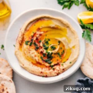 A top-down shot of a beautiful, perfectly smooth hummus in a white bowl, garnished with a swirl of olive oil, paprika, and fresh parsley. Served with pita chips on the side, ready to eat.