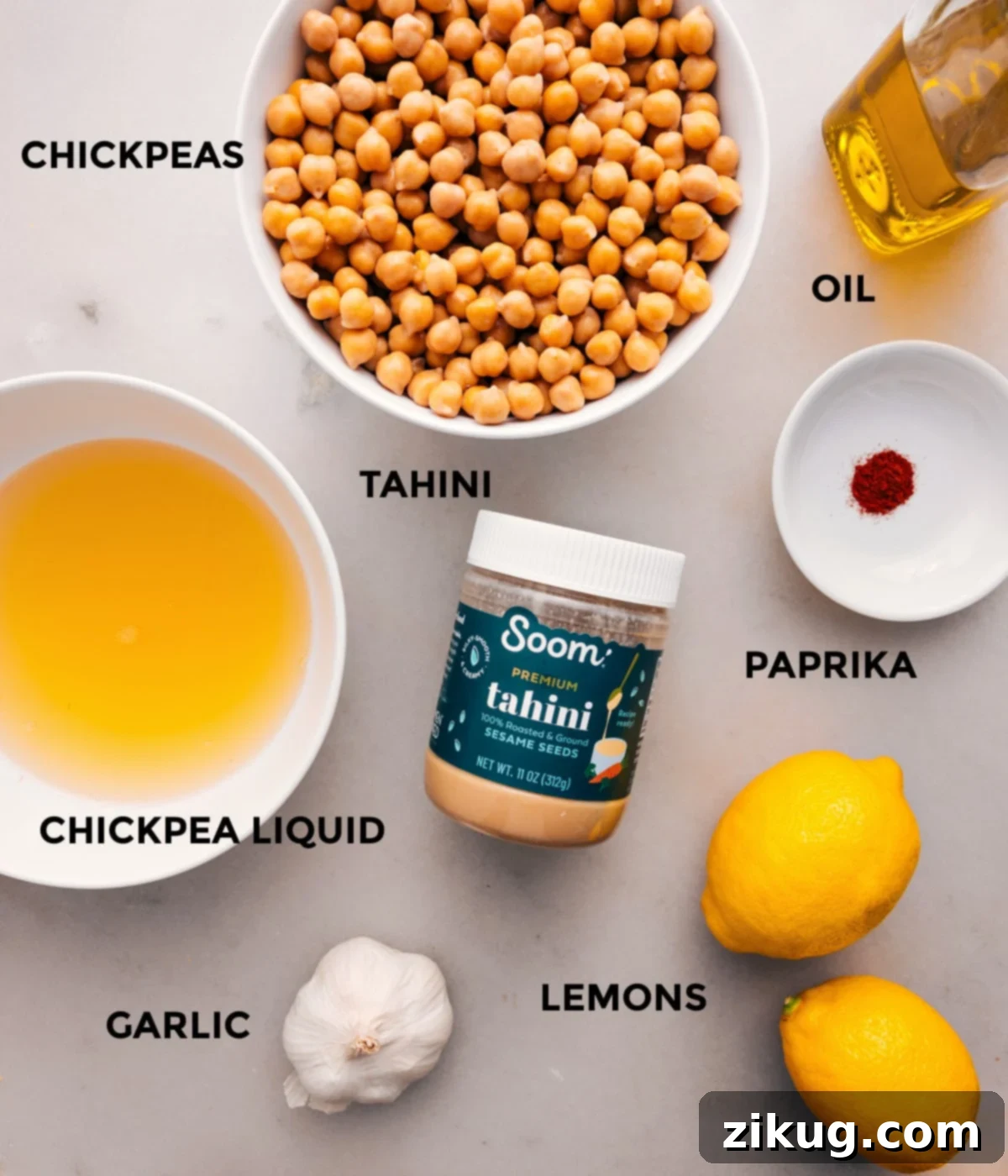 A vibrant display of all the essential ingredients prepped for making homemade hummus, including gleaming olive oil, canned chickpeas, rich tahini, a bowl of paprika, fresh lemons, and whole garlic cloves.