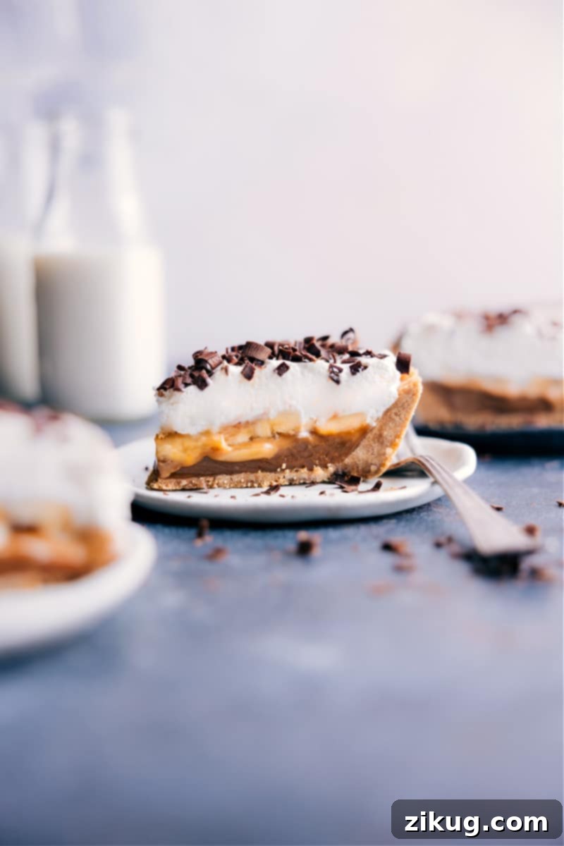 A delectable slice of Banoffee Pie on a white plate, showcasing its distinct layers of golden crust, rich caramel, sliced fresh bananas, and a generous topping of fluffy white whipped cream, garnished with chocolate shavings.