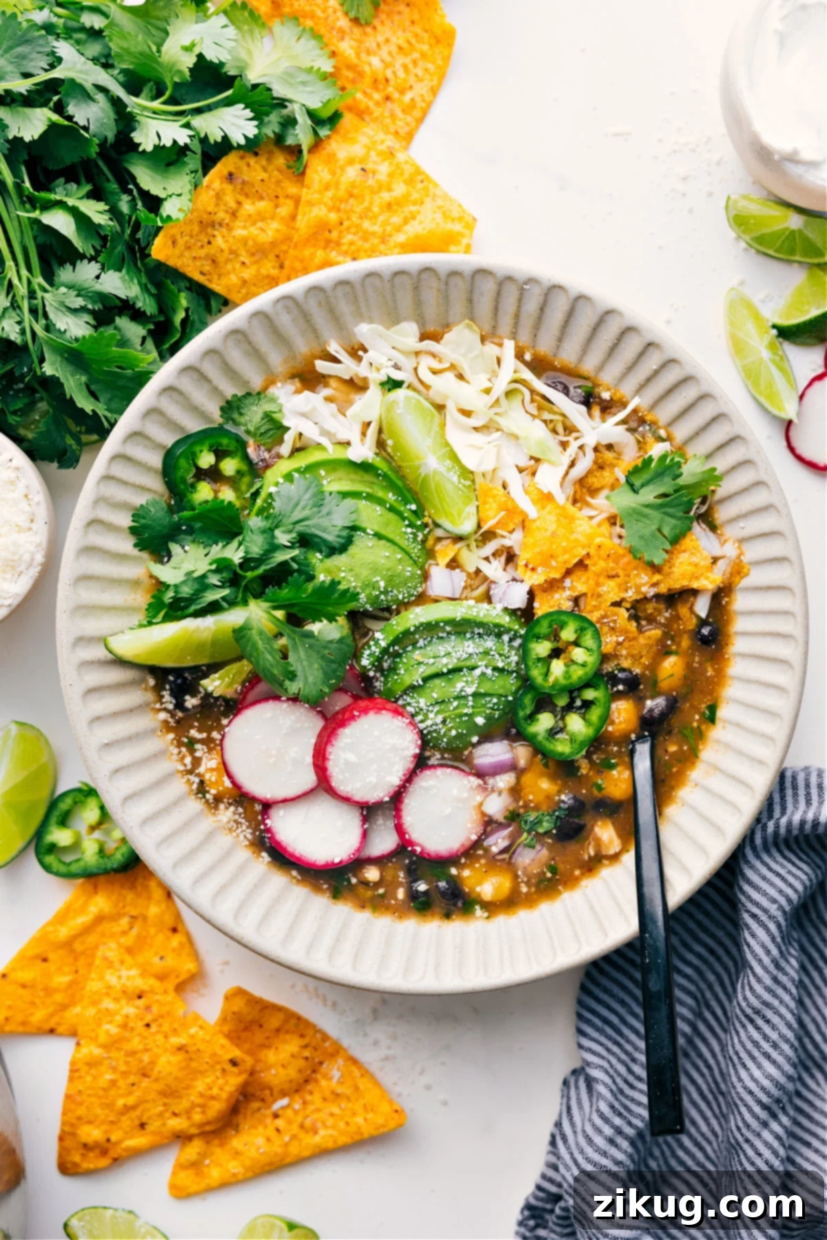 Quick & Easy Chicken Pozole Verde Recipe A steaming bowl of Quick Chicken Pozole Verde garnished with shredded cabbage, crispy tortilla chips, creamy avocado slices, thinly sliced radishes, fresh cilantro, zesty lime wedges, and a slice of jalapeño.