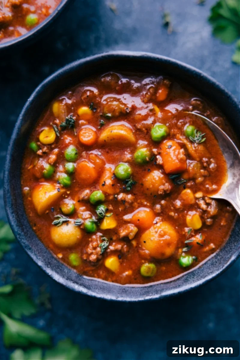 Overhead shot of a steaming bowl of homemade Vegetable Beef Soup, brimming with tender ground beef, carrots, celery, potatoes, peas, and corn in a rich, savory tomato-based broth, garnished with fresh parsley.