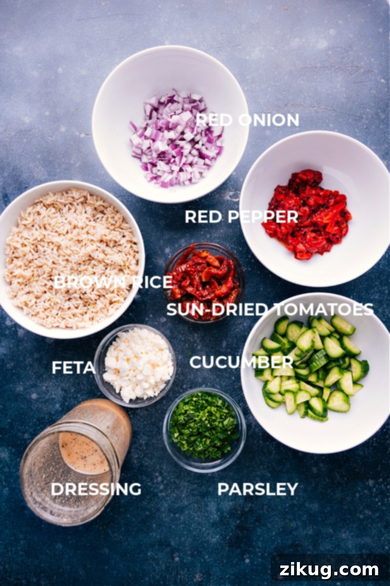 A flat lay image displaying all the fresh and colorful ingredients needed for the Brown Rice Salad, including cucumbers, roasted red peppers, sun-dried tomatoes, red onion, parsley, and feta.
