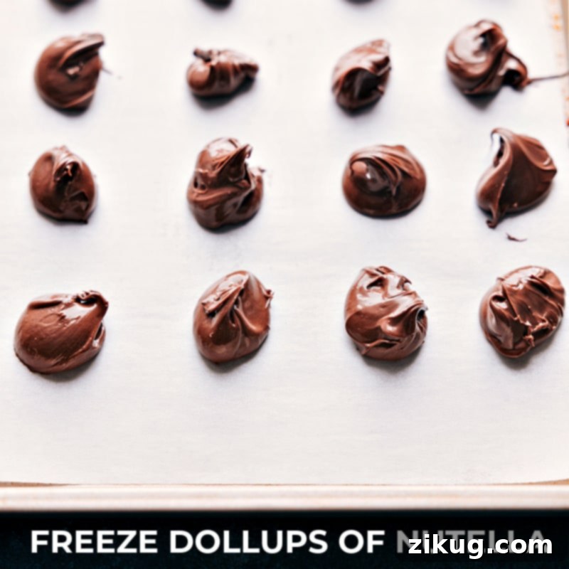 Prepare for the ultimate sweet indulgence: Nutella-Stuffed Chocolate-Chip Cookies! Enjoy a perfect blend of crunchy and chewy, packed with two kinds of chocolate chips and a delightful gooey Nutella core. Recipe via chelseasmessyapron #dessert #best #quick #easy #simple #familyfriendly #kidfriendly #comfortfood #nutellastuffed #chocolatechip #cookies Step-by-step images showing dollops of Nutella being placed on a parchment-lined sheet pan for freezing