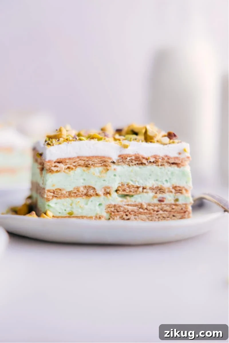 A close-up image of a perfect slice of Pistachio Icebox Cake, showing the distinct layers of graham crackers, green pistachio pudding, and white whipped cream, with a light sprinkle of chopped pistachios.