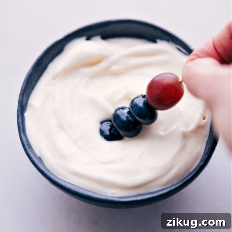 A hand dipping a fresh strawberry into the creamy white fruit dip on the rainbow platter