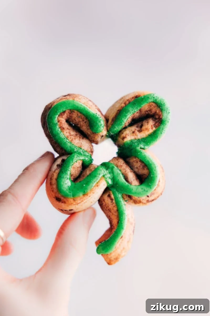 St. Patrick's Day is just around the corner and what better way to celebrate than with some delicious Shamrock Cinnamon Rolls? These adorable treats are not only festive and fun, but they're also incredibly easy to make. With just a can of refrigerated cinnamon rolls and some green food coloring, you can transform a classic breakfast pastry into a holiday masterpiece that kids and adults alike will love! Recipe via chelseasmessyapron #dessert #breakfast #quick #easy #simple #brunch #stpatricks #stpatricksday #shamrock #cinnamon #roll Shamrock cinnamon roll being held up, showcasing its green frosting and festive shape.