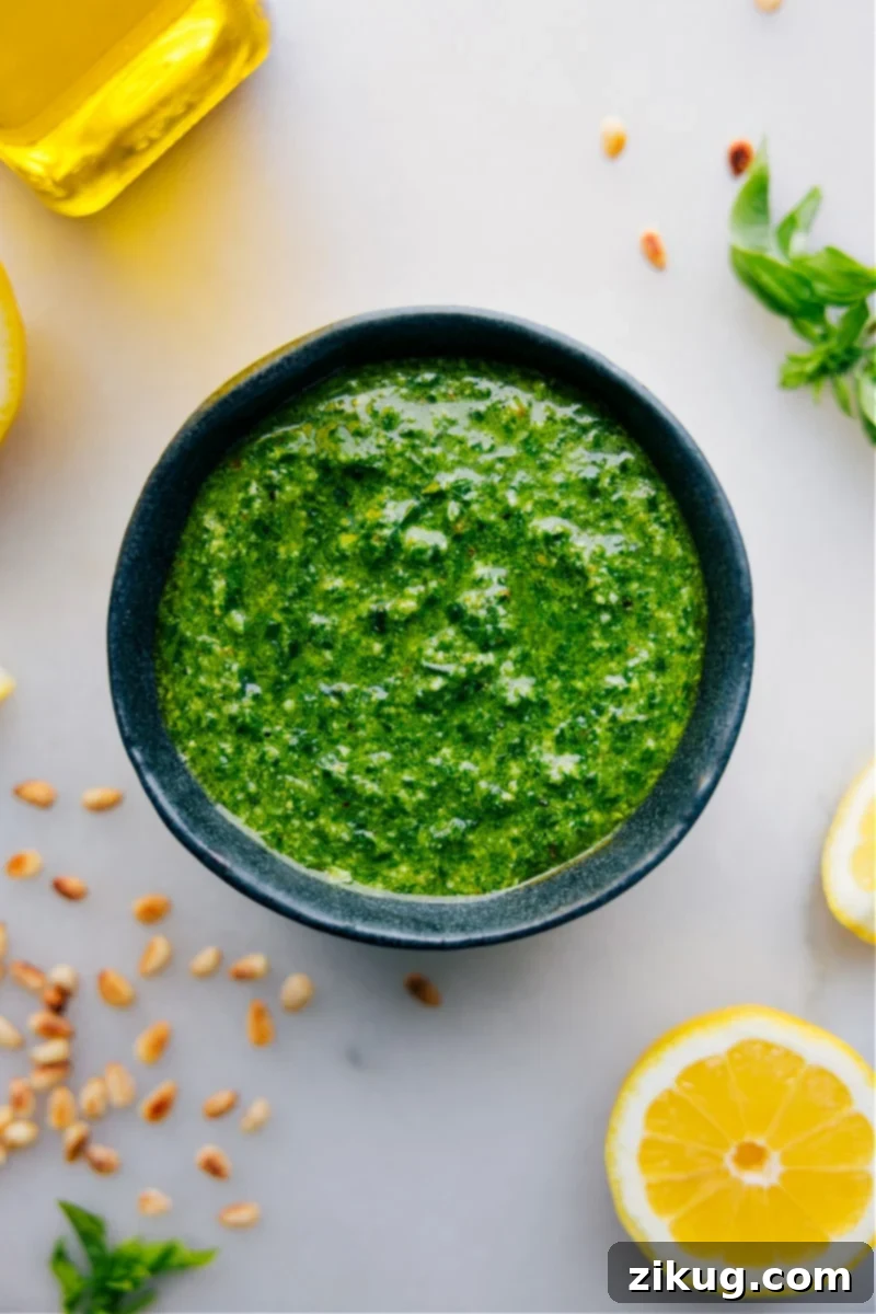 Overhead image of freshly made Pesto sauce in a bowl with basil leaves, pine nuts, and Parmesan around it