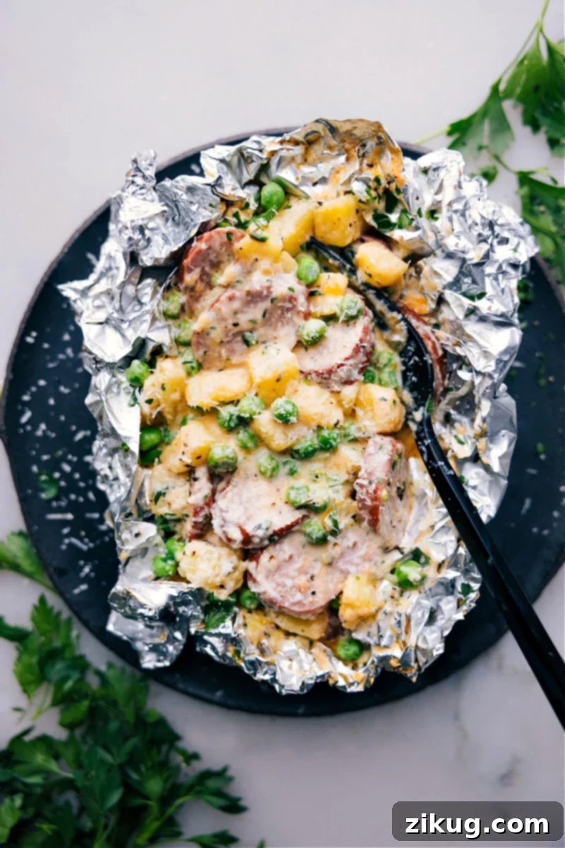 Creamy Sausage and Potato Foil Packs are a simple and delicious dinner option that's perfect for busy weeknights or camping trips. These foil packs are loaded with savory smoked sausage, tender golden potatoes, and sweet peas, all smothered in a rich, creamy, and cheesy sauce. The best part? They are incredibly easy to make and require minimal cleanup! Recipe via chelseasmessyapron #dinner #quick #easy #simple #familyfriendly #kidfriendly #campfiremeals #campingmeals #comfortfood #creamy #sausage #potato #foilpacks Overhead shot of Creamy Sausage and Potato Foil Packs, a hearty and convenient meal for families and outdoor enthusiasts.