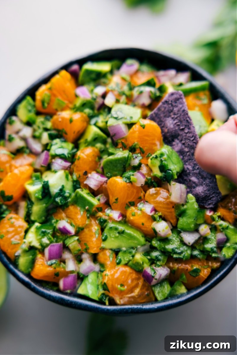 A close-up of Avocado-Orange Salsa in a rustic bowl, nestled amongst fresh ingredients like whole oranges, avocados, and cilantro sprigs, on a wooden board.
