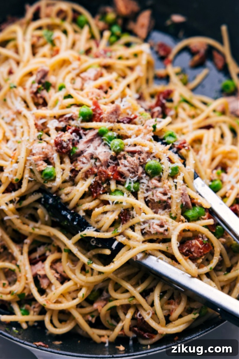 Serving Healthy Tuna Salad Pasta with Tongs Close-up shot of tongs expertly scooping a generous portion of healthy tuna pasta from a large serving bowl, highlighting the perfectly coated noodles, vibrant peas, and flaky tuna, ready for plating and garnished with freshly grated Parmesan cheese.