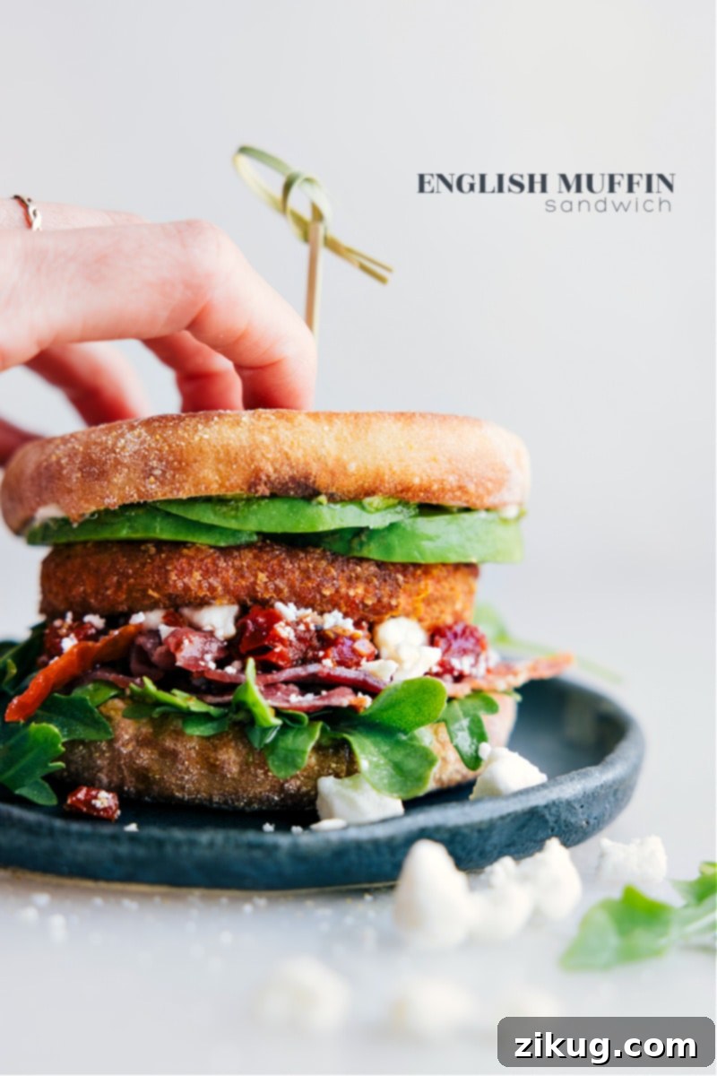 The Ultimate English Muffin Sandwich: Avocado, Pesto, Feta, Bacon, Sweet Potato Hash Brown, Sun-Dried Tomatoes & Greens A perfectly assembled English Muffin Sandwich on a white plate, showcasing layers of avocado, pesto, feta, bacon, and a crispy sweet potato hash brown, with sun-dried tomatoes and fresh greens peeking out.
