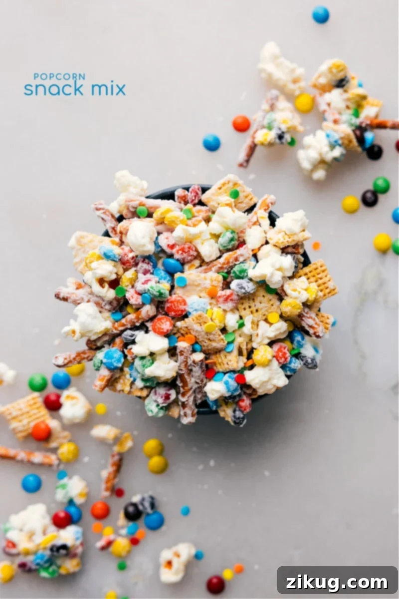 This Popcorn Snack Mix is a perfect blend of sweet and salty, featuring popcorn, pretzels, Chex cereal, and mini M&M's coated in white chocolate and sprinkles. An easy, no-bake recipe for any sweet craving or gathering. Overhead image of the Popcorn Snack Mix ready to be enjoyed, showcasing popcorn, pretzels, Chex, M&M's, white chocolate, and sprinkles.