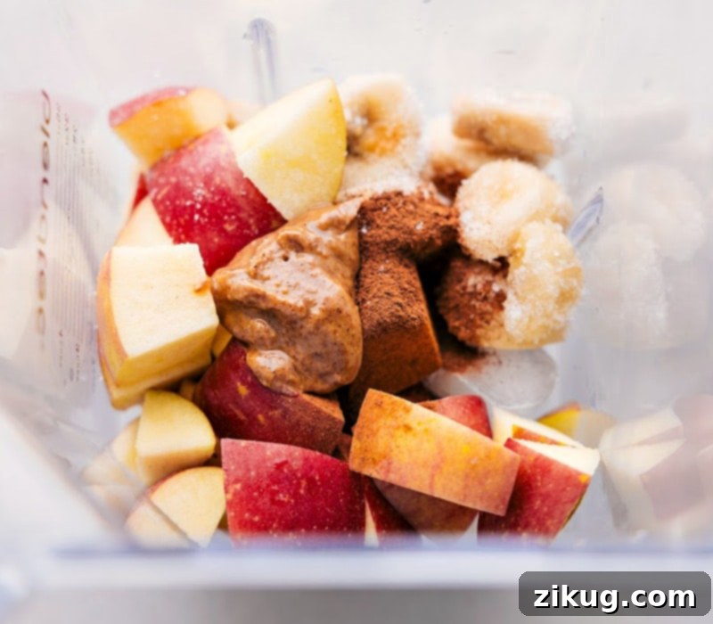 Various ingredients for an Apple Pie Smoothie being dropped into a blender: sliced apples, a banana, a spoonful of almond butter, and a sprinkle of spices. The blender is half-full, ready for processing.