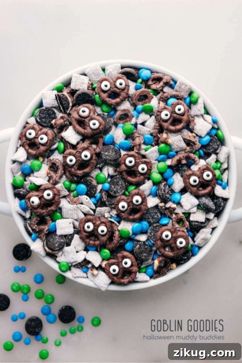 These Goblin Goodies (Halloween Muddy Buddies) are a fun Halloween treat, bursting with rich flavors of chocolate and peanut butter, complemented by the crunch of cereal, Oreos, and M&M's. It's a spooky and delicious snack mix you won't want to miss! Recipes via ChelseasMessyApron.com #halloween #oreos #M&Ms #chex #muddybuddies #snackmix Overhead view of a bowl of Goblin Goodies (Halloween Muddy Buddies) featuring chocolate and peanut butter coated cereal, mini Oreos, blue and green M&M's, and spooky pretzel goblins with edible eyes.
