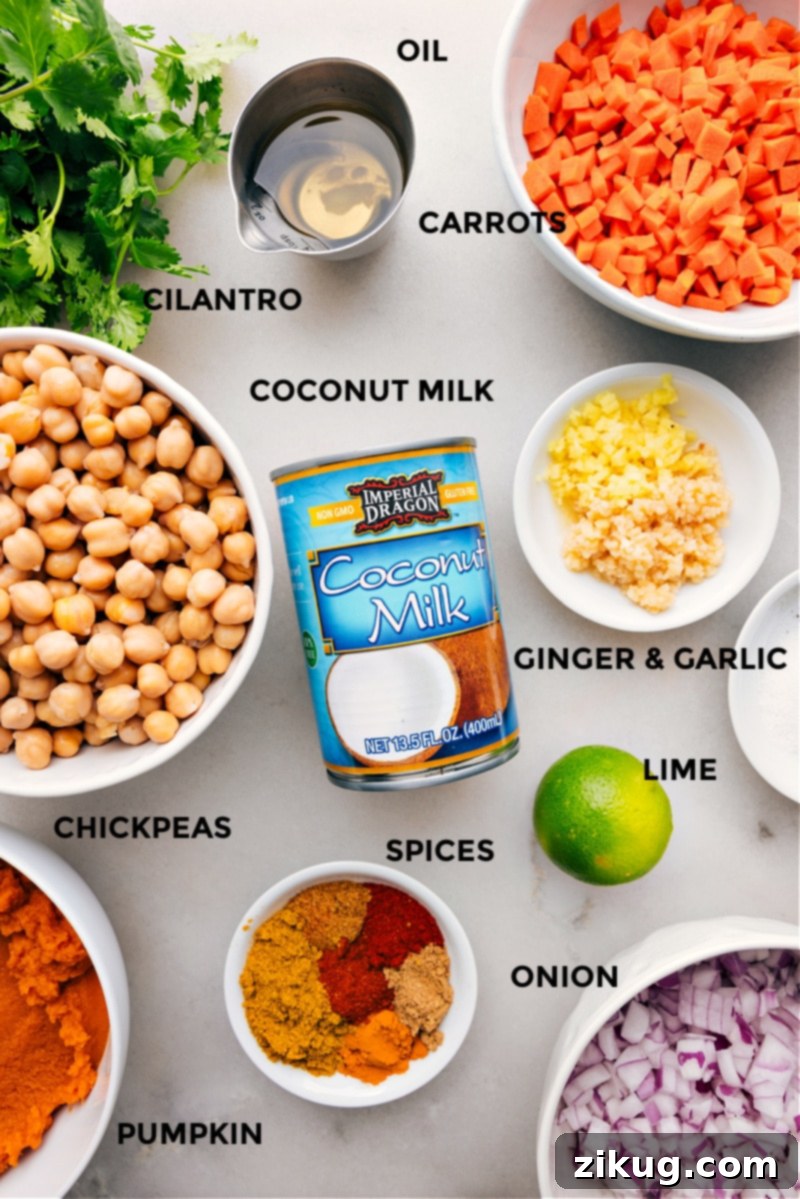 A flat lay photograph showcasing all the fresh and canned ingredients needed to make a delicious Pumpkin Curry: red onion, carrots, garlic, ginger, full-fat coconut milk, canned pumpkin puree, chickpeas, and a selection of spices.