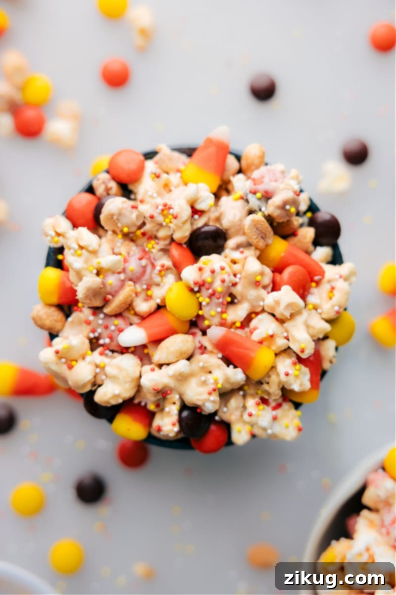 Candy Corn Popcorn: An addictive sweet and salty fall snack mix with peanut butter, white chocolate, and vibrant candy corn. Ideal for Halloween treats and kid-friendly parties. Recipe via ChelseasMessyApron.com #candycorn #popcorn #whitechocolate #peanutbutter #halloween #treat #kidfavorite Overhead view of a large bowl overflowing with vibrant Candy Corn Popcorn, featuring fluffy white popcorn beautifully coated in a creamy mixture, interspersed with yellow, orange, and white candy corn pieces, glistening white chocolate, and crunchy dry-roasted peanuts. The inviting mix sits on a rustic wooden surface, perfect for a festive fall or Halloween celebration.