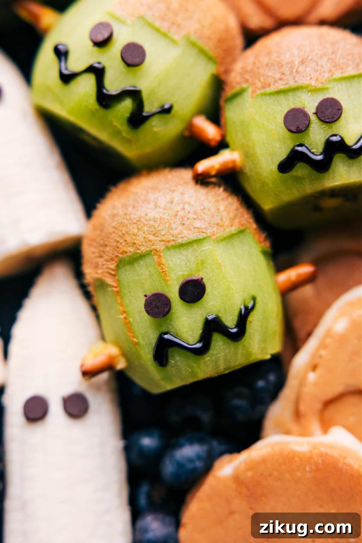 A creative Halloween treat featuring a kiwi carved and decorated to resemble Frankenstein's face, complete with pretzel bolts and chocolate chip eyes.