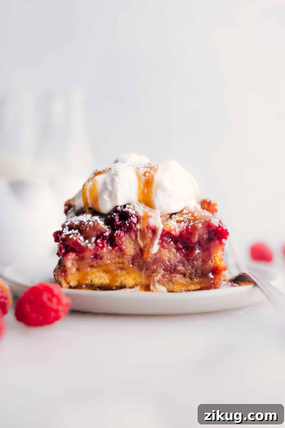 A delectable slice of Raspberry Bread Pudding on a plate, generously adorned with whipped cream and a drizzle of vanilla sauce, ready to be savored.
