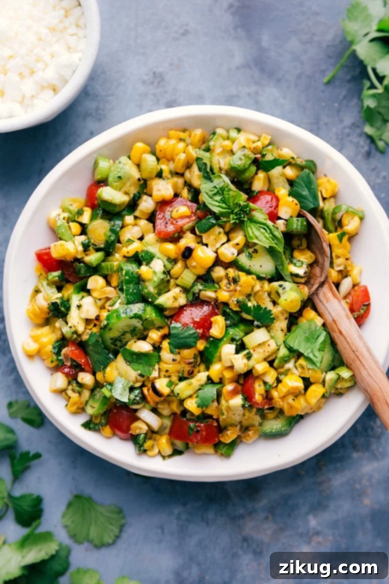 A large bowl brimming with vibrant, fresh corn salad, featuring golden corn kernels, red cherry tomatoes, green herbs, and white feta cheese, with a serving spoon ready to dish out. The salad is beautifully presented and ready to be enjoyed.